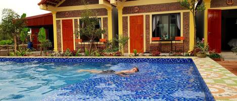 Piscina com azulejos de mosaico, cercada por um edifício com portas numeradas e um caminho revestido de azulejos.