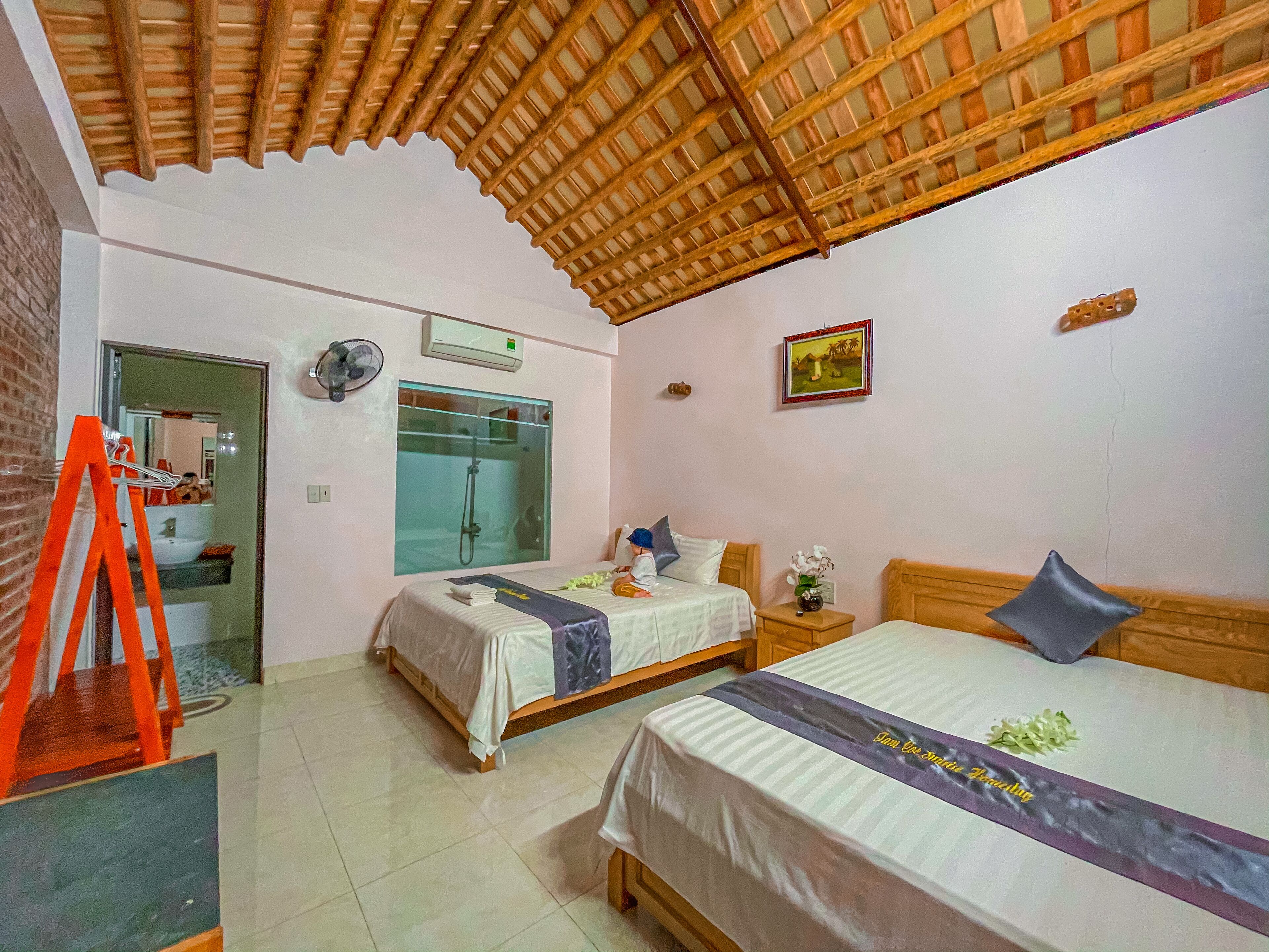 Habitación de hotel con dos camas, techo de madera, ducha de vidrio, ventilador y una pequeña mesa con un florero.