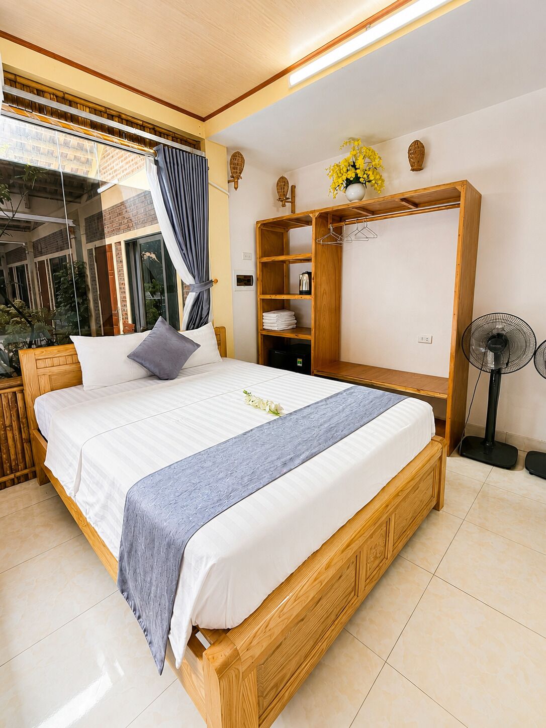 A bedroom with a bed, a wooden wardrobe, a fan, and a window with curtains.