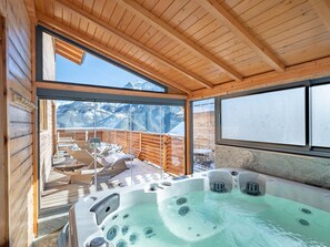 A wooden cabin with a hot tub, wooden deck, and a view of snow-covered mountains.