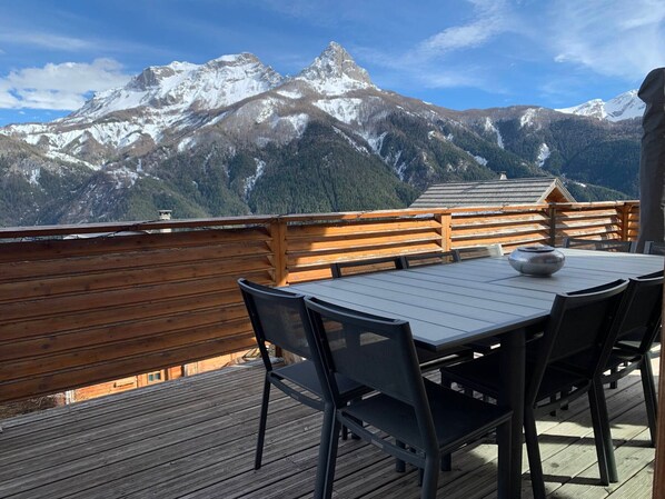 A wooden deck with a table and chairs, overlooking snow-capped mountains.