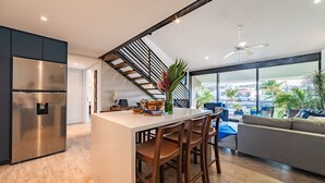 Une cuisine moderne dotée d’un réfrigérateur en acier inoxydable, d’un coin bar avec des tabourets et d’un escalier menant à un balcon offrant une vue sur les palmiers.