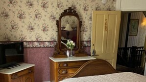 A bedroom with a wooden dresser, a television on a stand, a bed with a white comforter, a floral wallpaper, and a door with a nameplate. - AH #15 · AH #15 Queen w/Shower (Wabasha)