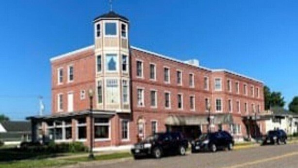 A two-story brick building with a central tower, a covered entrance, and a parking area with cars. - AH #15 · AH #15 Queen w/Shower (Wabasha)