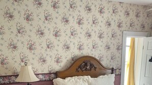 A bedroom with a wooden bed, a red chair, a nightstand with a lamp, and floral wallpaper. - AH #15 · AH #15 Queen w/Shower (Wabasha)