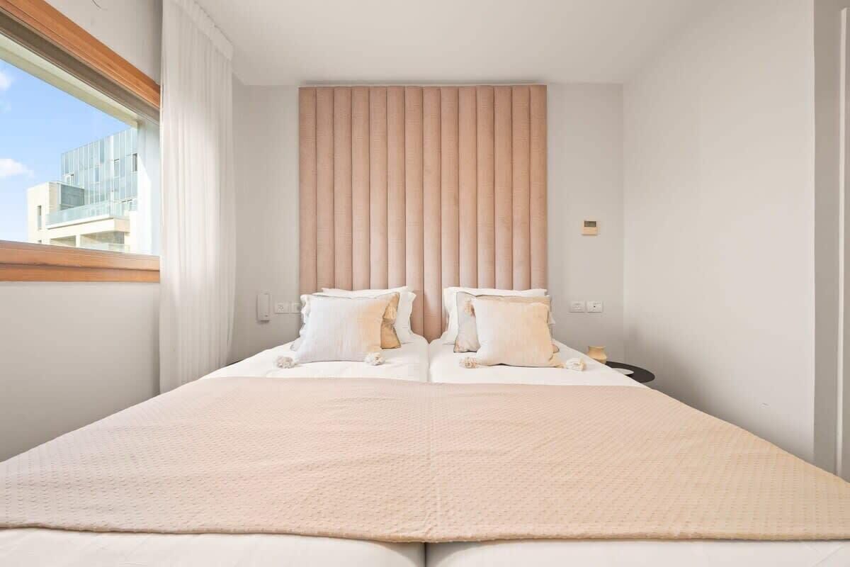 A modern hotel room with a large bed, a window with blinds, and a view of a building outside.
