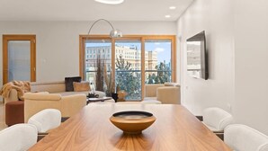 A modern living room with a wooden dining table, white armchairs, a sofa, and large windows offering a city view.