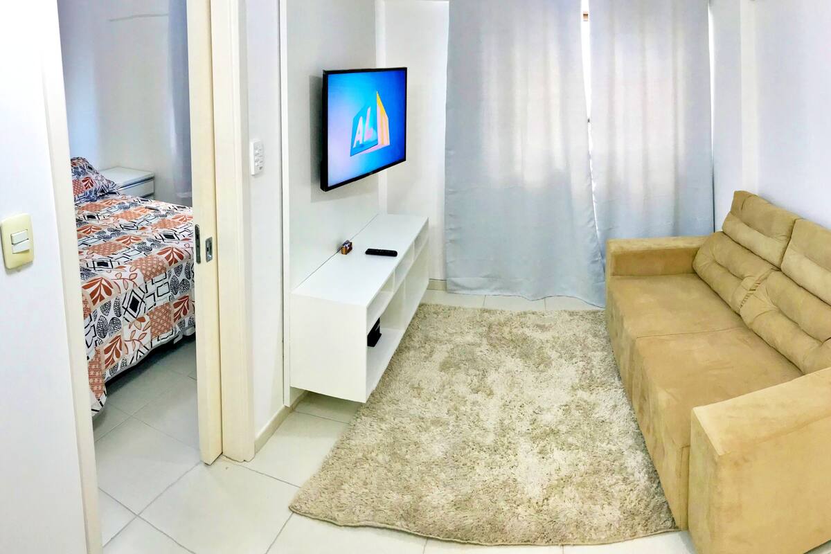 A living room with a beige sofa, a TV on a stand, a patterned bed, and a small white cabinet.