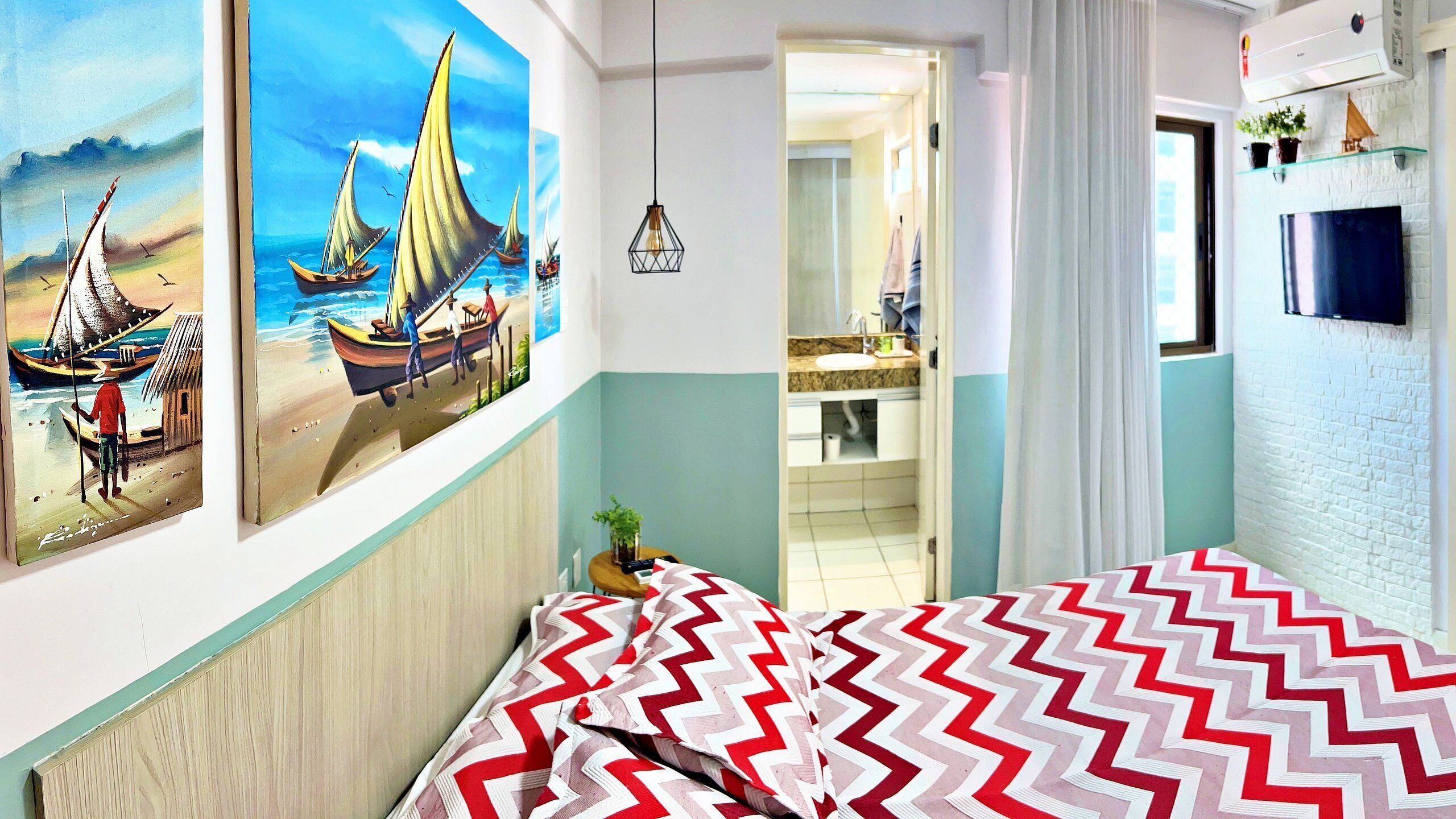 A bedroom with a bed, a painting of boats, a bathroom with a mirror, and a TV mounted on the wall.