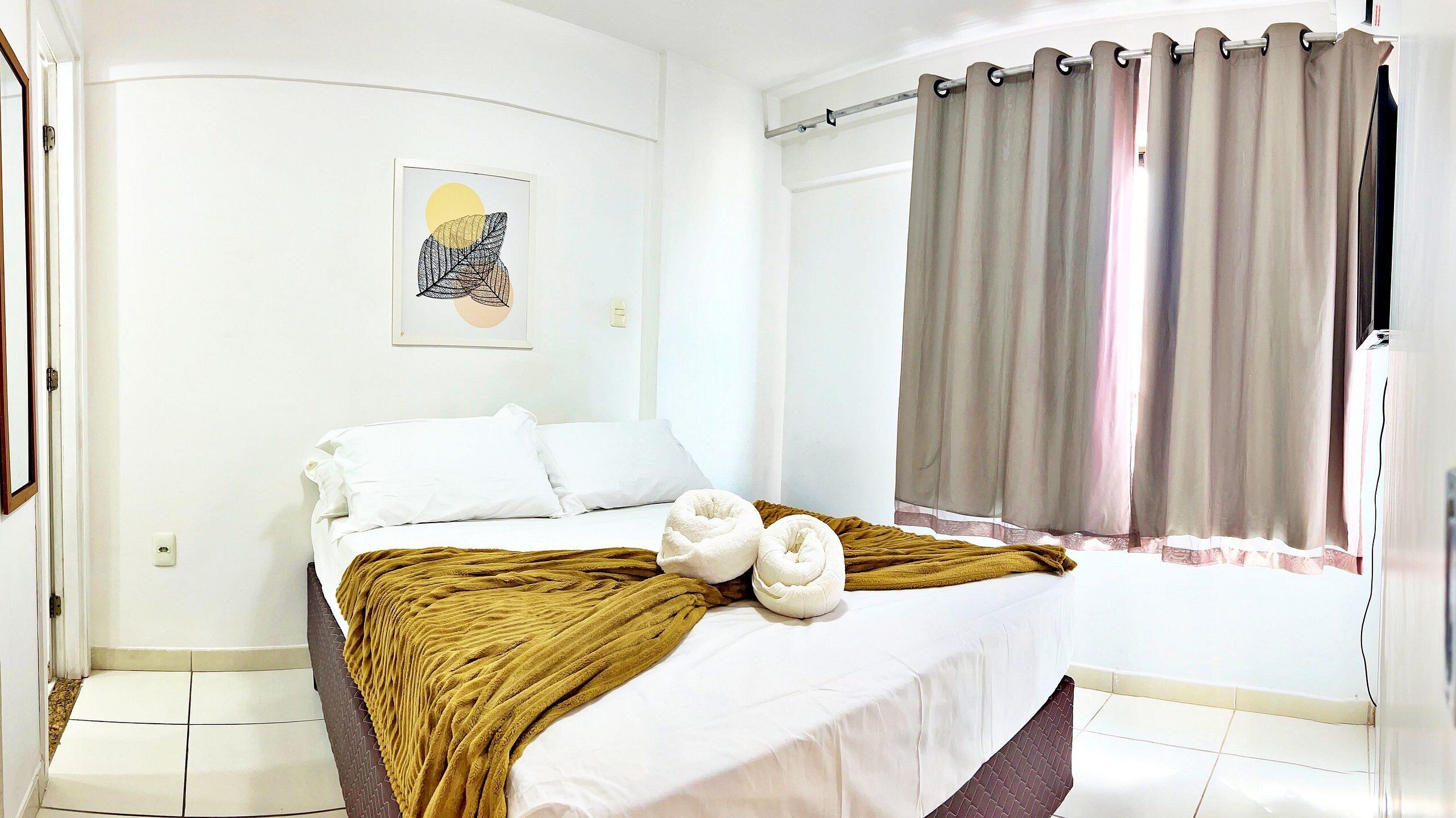 A bedroom with a bed, a window with curtains, a wall-mounted light switch, and a framed picture.