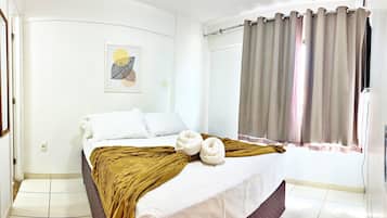 A bedroom with a bed, a window with curtains, a wall-mounted light switch, and a framed picture.