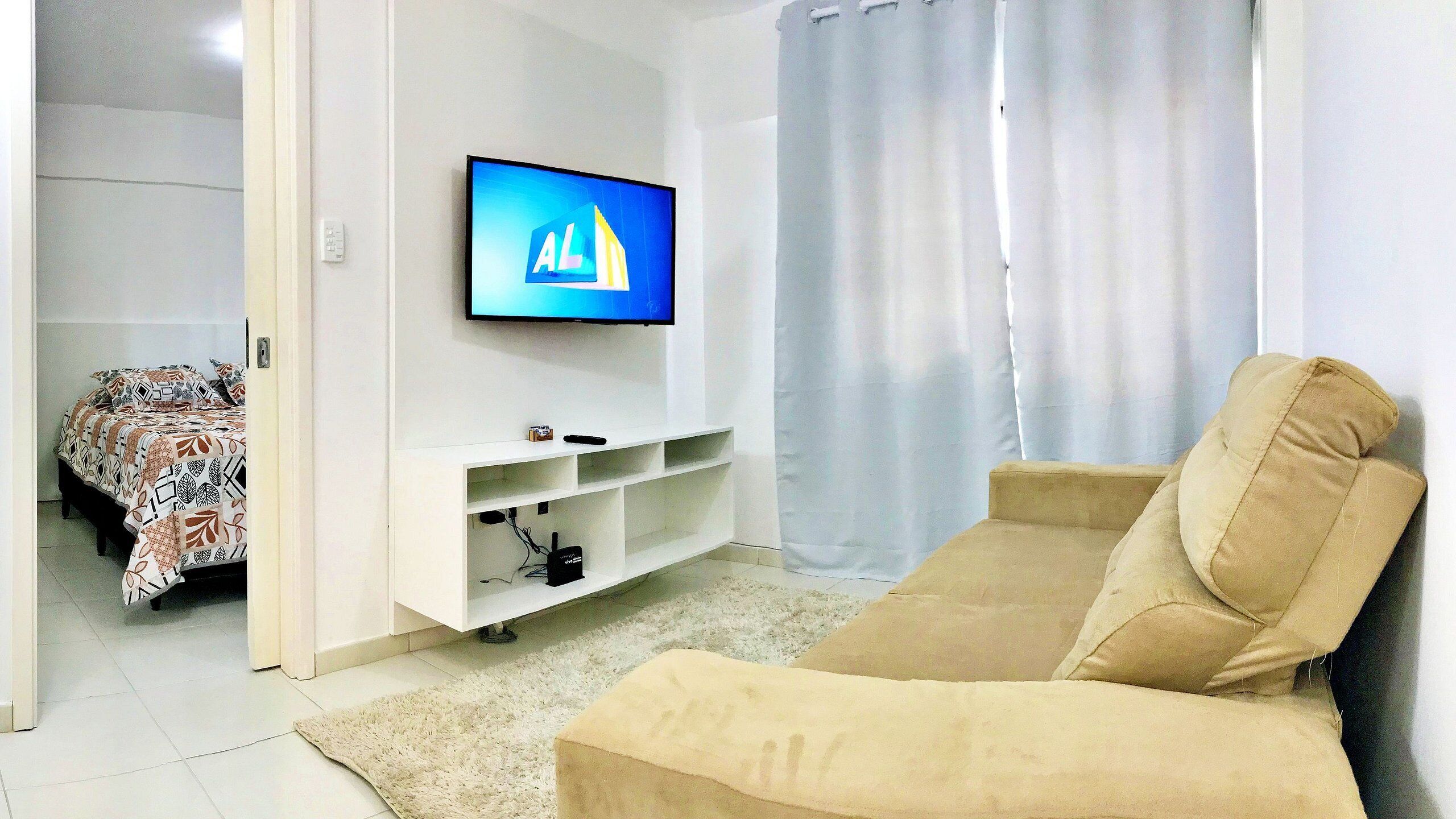 A living room with a beige sofa, a flat-screen TV mounted on the wall, a white entertainment unit below, and a patterned bed in the background.