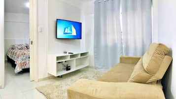 A living room with a beige sofa, a flat-screen TV mounted on the wall, a white entertainment unit below, and a patterned bed in the background.