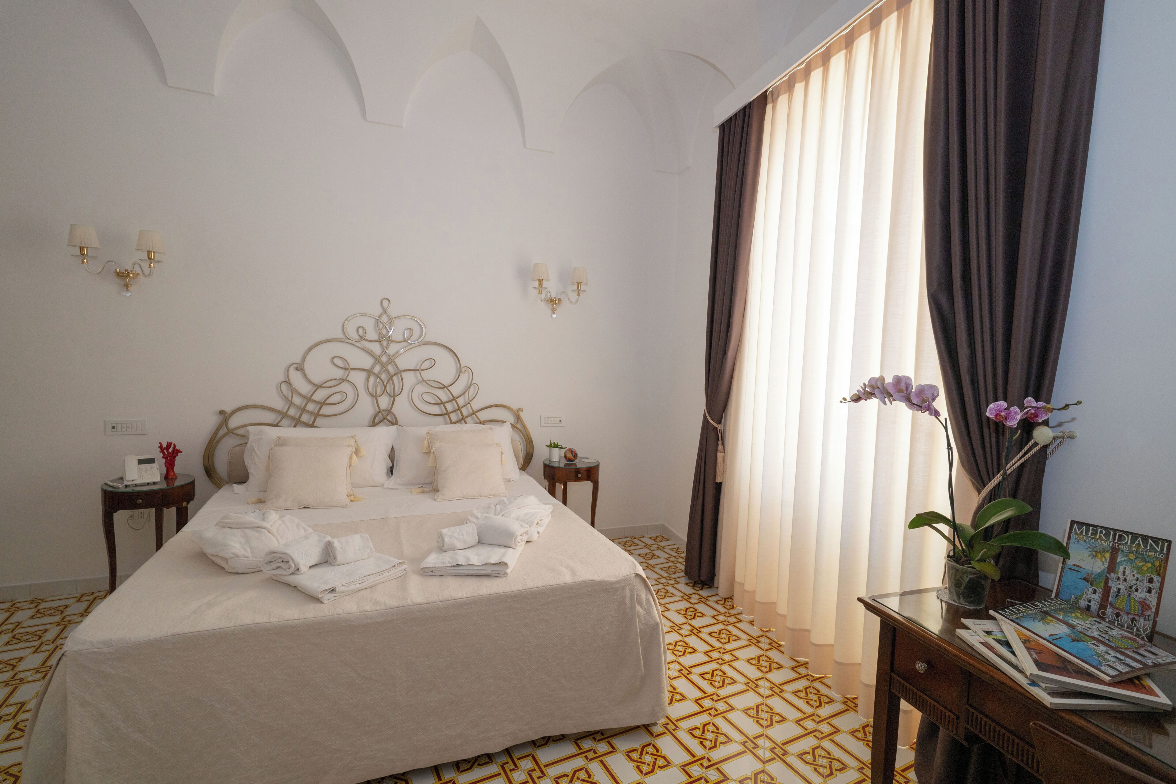 Quarto de hotel com uma cama grande, duas mesas de cabeceira, uma luminária fixada na parede, uma janela com cortinas e uma pequena mesa com uma revista.