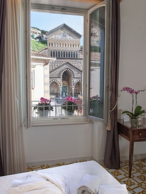A hotel room with a bed, a window with curtains, a bedside table with a plant, and a view of a historic building. - Core Amalfitano City Suites (Amalfi)