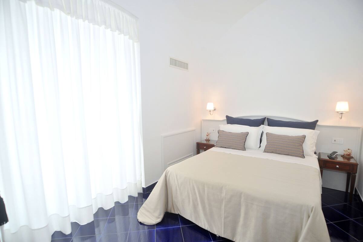 A neatly made bed with a white bedspread and two pillows, flanked by nightstands with lamps, in a room with white walls and a large window with sheer curtains.