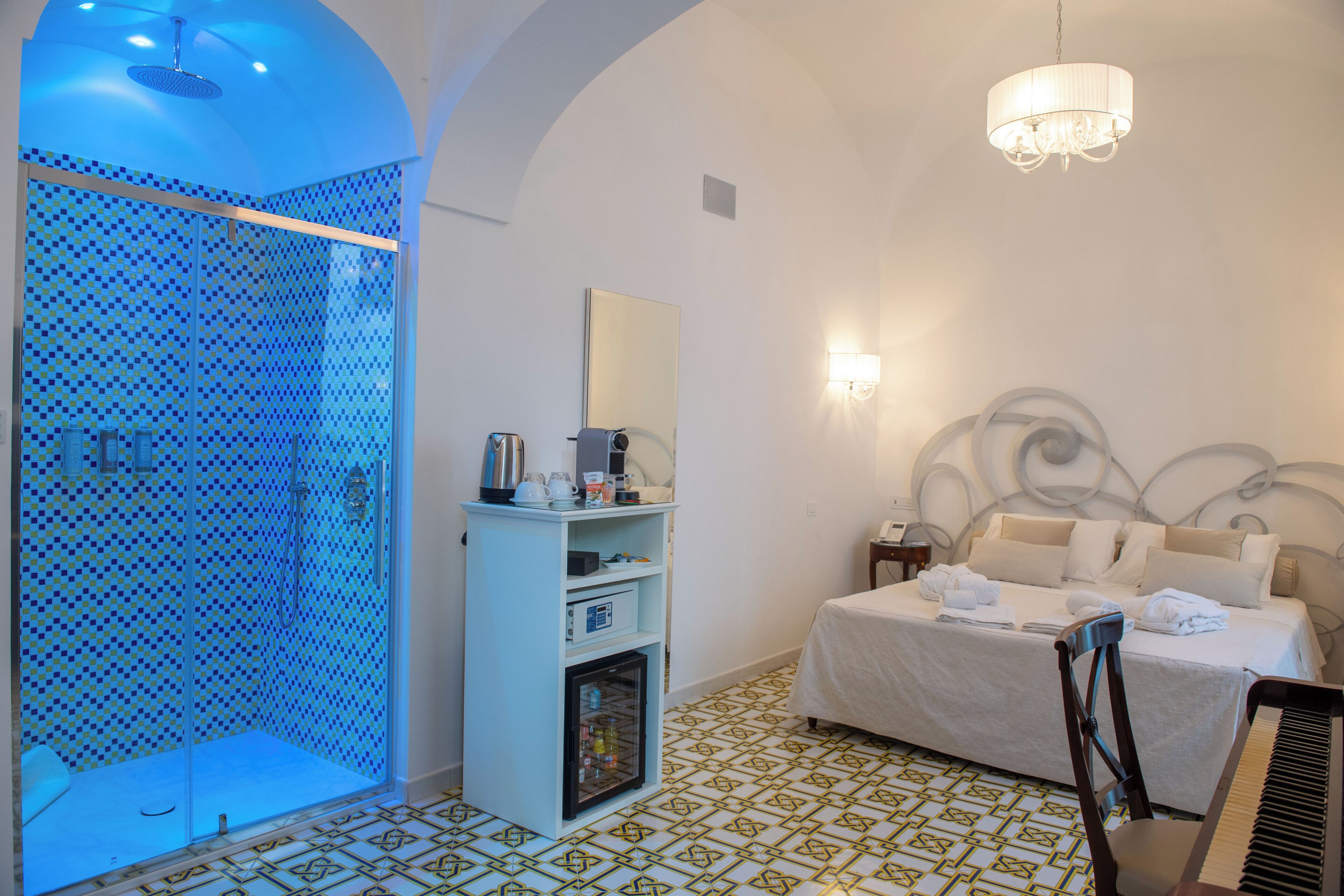 Quarto de hotel com chuveiro, cama, mesa de cabeceira, espelho e lustre.