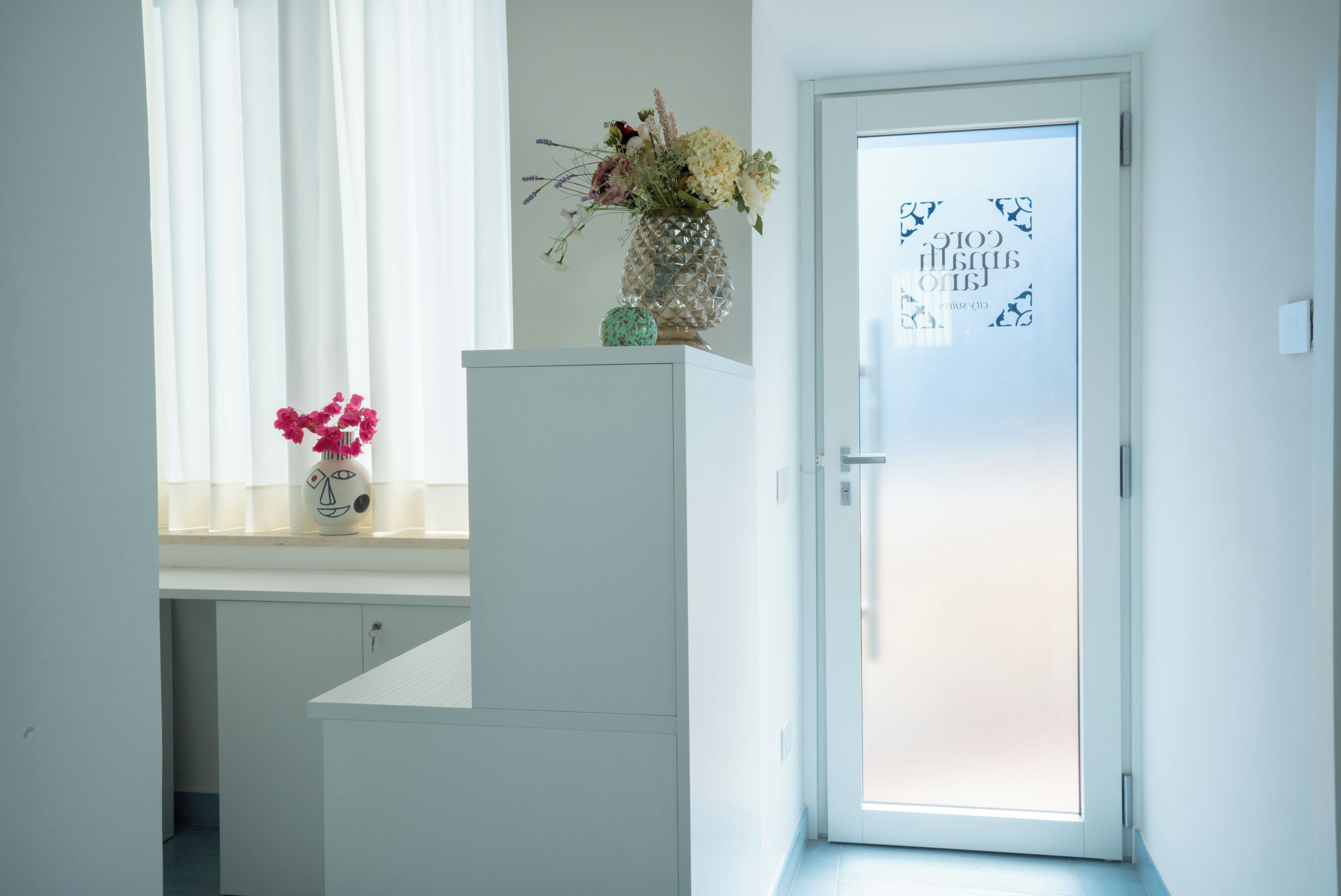 Una stanza con una porta bianca, una finestra con tende leggere, un vaso con fiori su un ripiano e un tavolino con un oggetto decorativo.