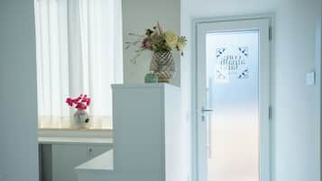 Una stanza con una porta bianca, una finestra con tende leggere, un vaso con fiori su un ripiano e un tavolino con un oggetto decorativo.