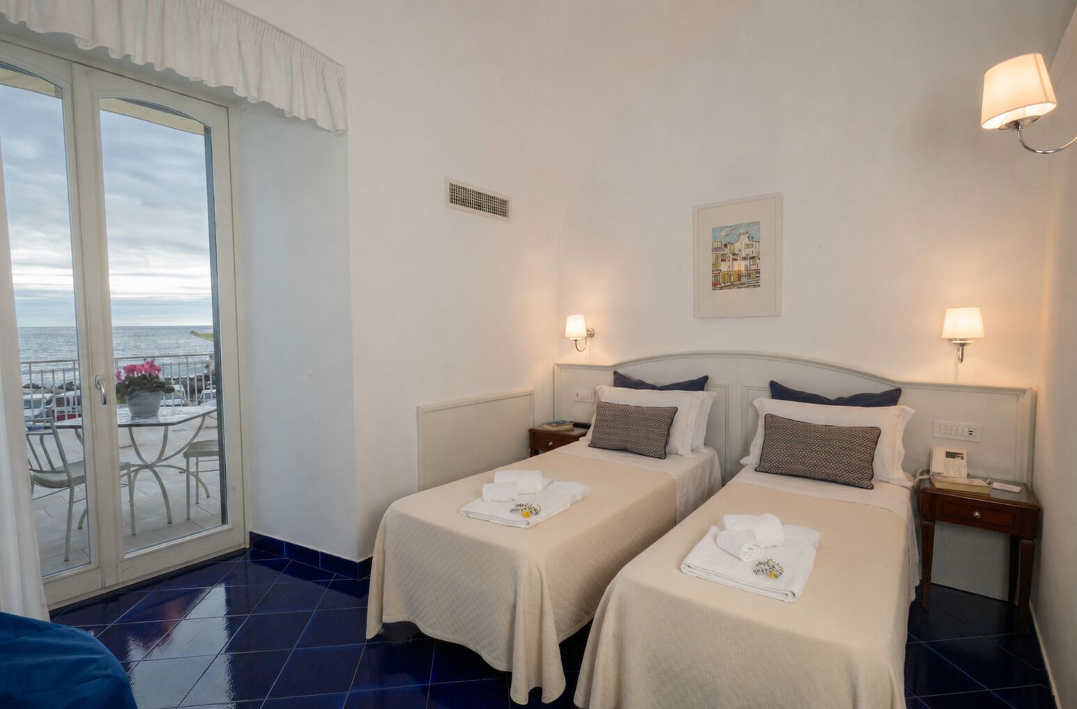 Una camera d'albergo con due letti, un balcone vista mare e un piccolo tavolo con sedie.