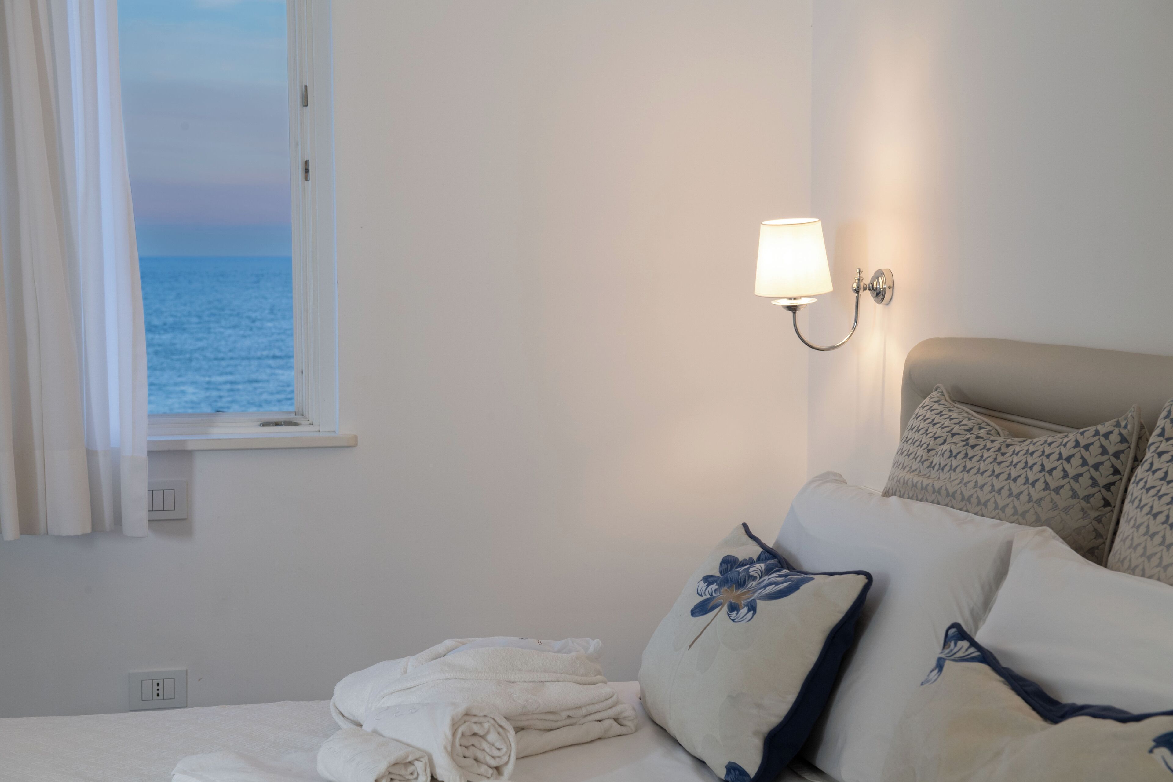 Una camera d'hotel con un letto, un comodino, una lampada, una finestra con tende e vista sul mare.