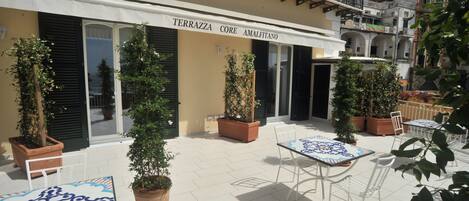 Un área de café al aire libre con mesas y sillas, plantas en macetas y un edificio con un cartel que dice