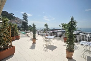 A terrace with potted plants, white metal chairs, and tables, overlooking a marina with boats and a coastal town in the background. - Core Amalfitano City Suites (Amalfi)