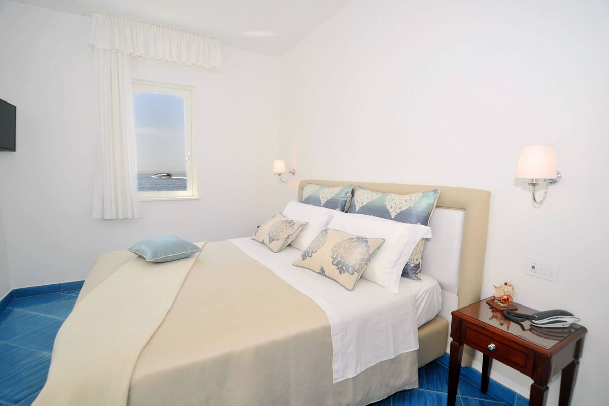 A bedroom with a bed, pillows, a nightstand, a lamp, a window with a view of the sea, and a blue floor.