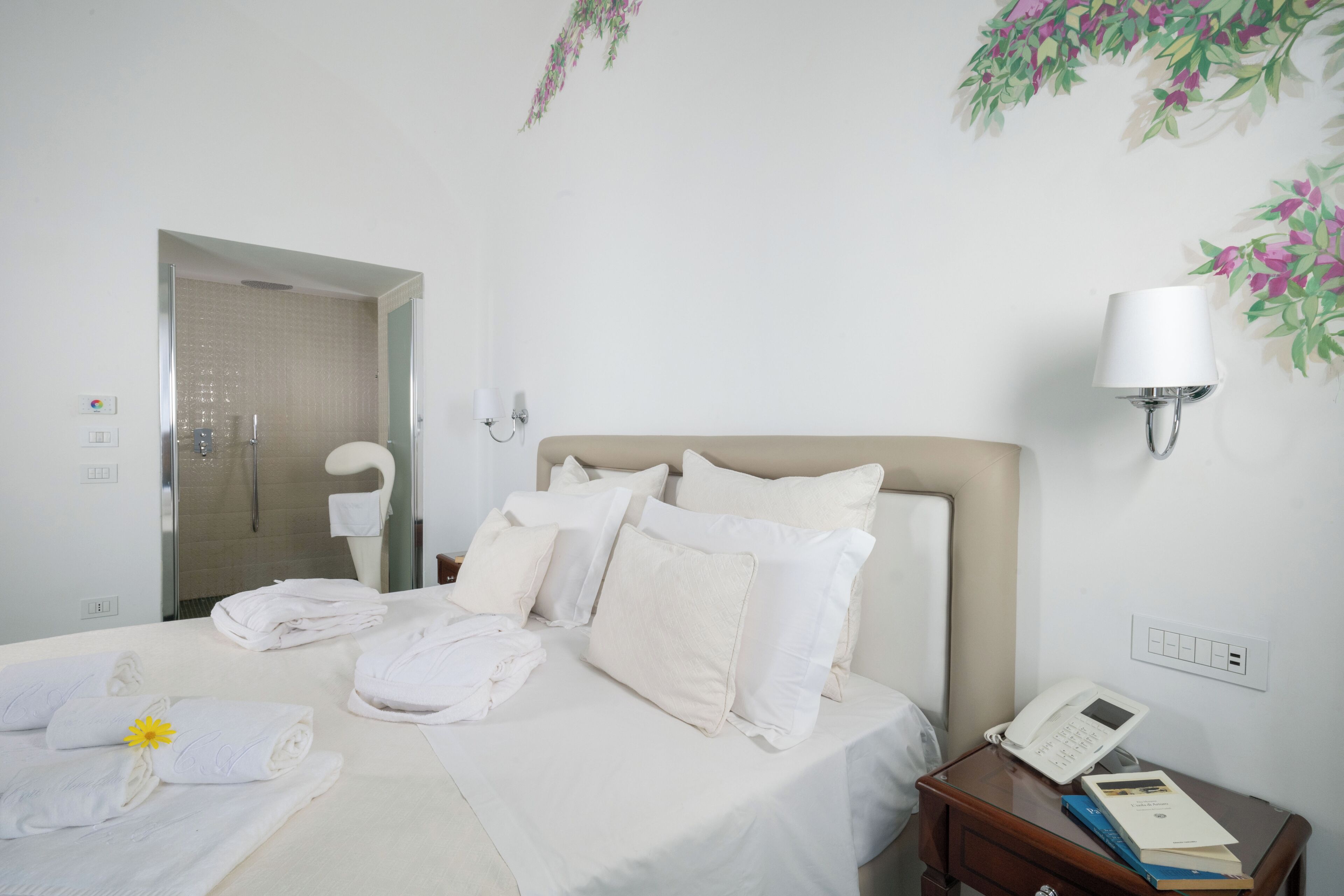 A hotel room with a bed, a bedside table with a phone and books, a wall-mounted lamp, and a shower area.
