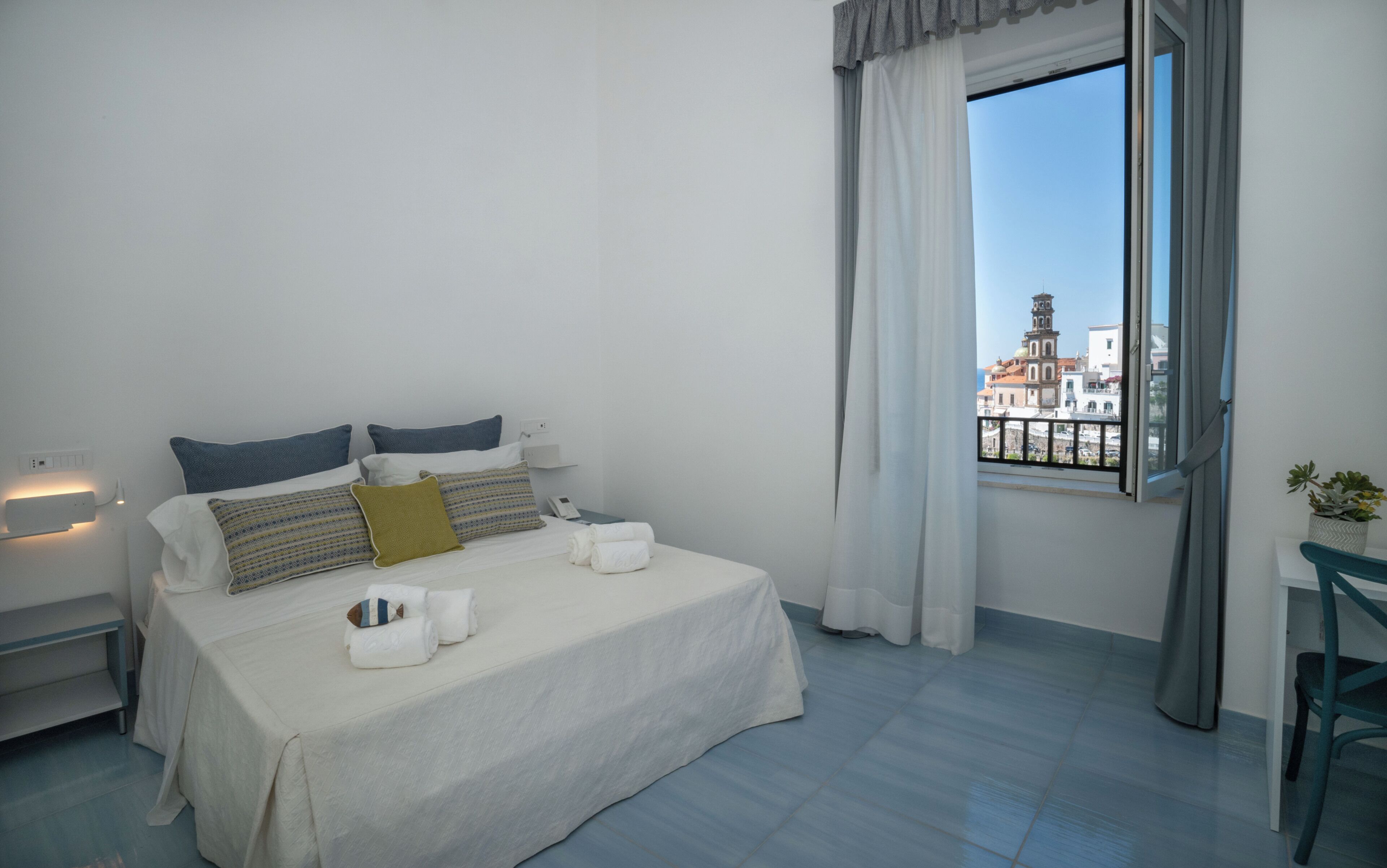 Una camera d'albergo con un letto, un comodino, una finestra che si affaccia su edifici e una tenda.