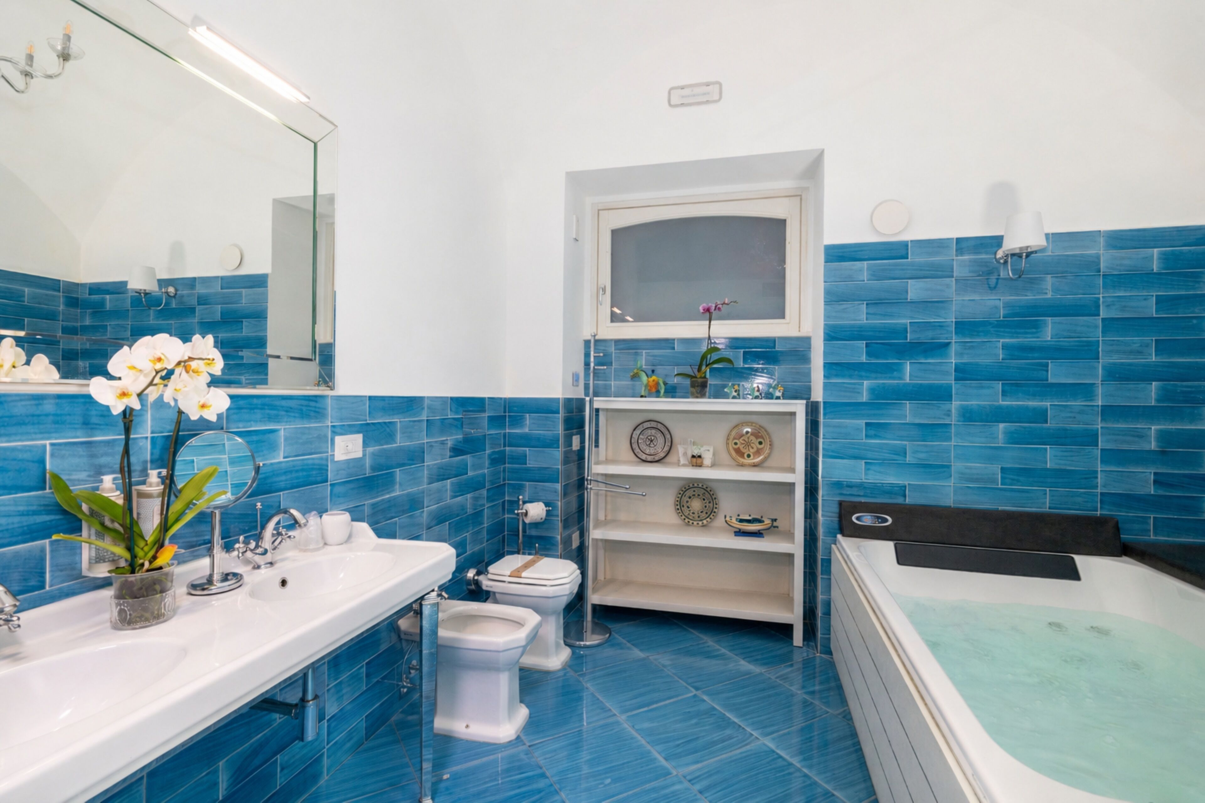 Un bagno con un lavandino bianco, due servizi igienici, una vasca da bagno e uno scaffale con oggetti decorativi.