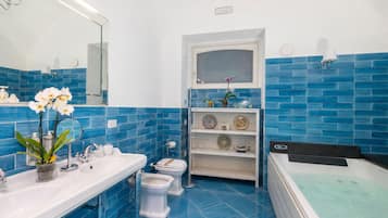 Un bagno con un lavandino bianco, due servizi igienici, una vasca da bagno e uno scaffale con oggetti decorativi.