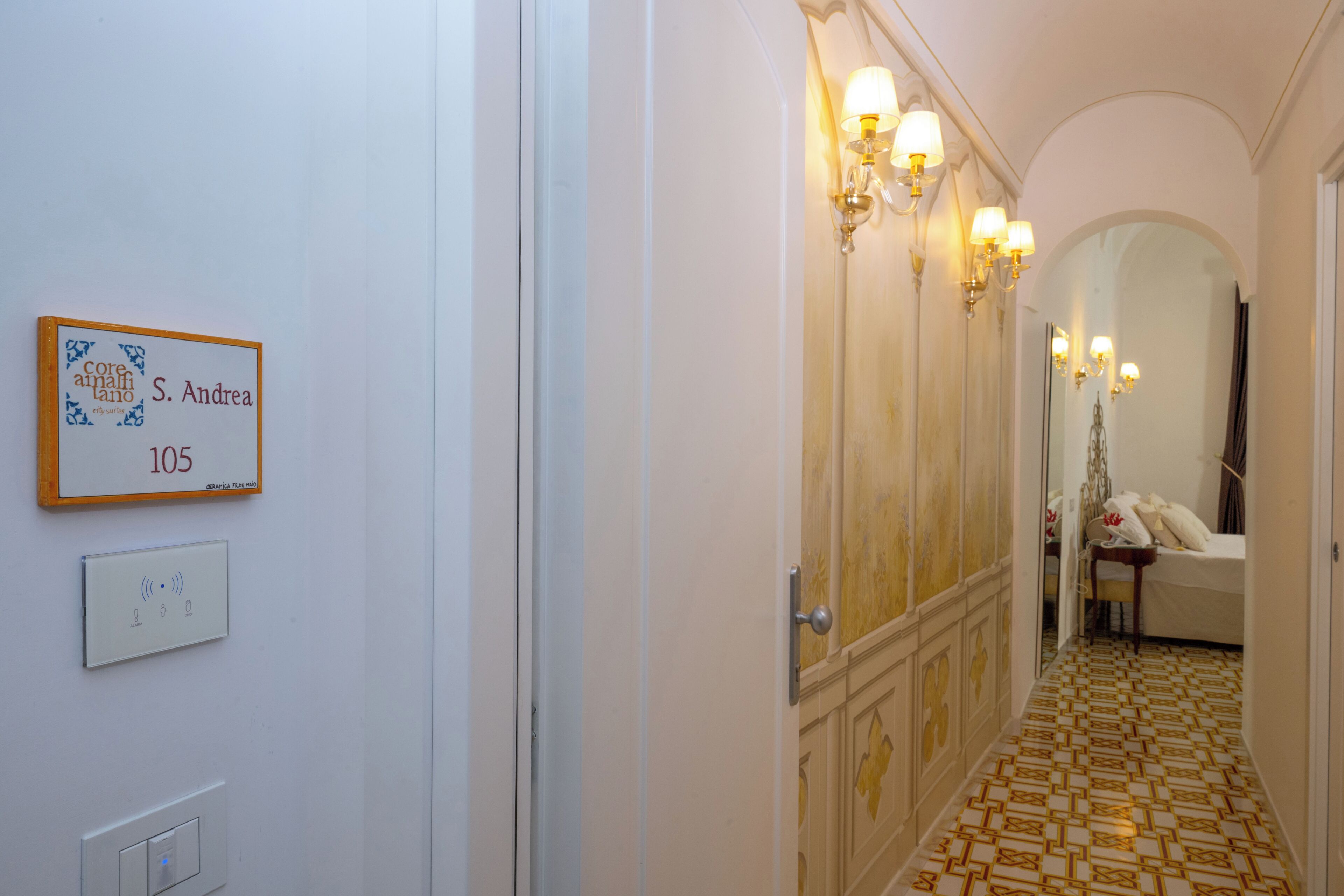 Un corridoio con una targa che indica la stanza numero 105, un interruttore della luce e una porta che conduce a una stanza con un letto e un tavolo.