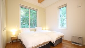 Un dormitorio con dos camas, una mesita de noche con lámpara, un calefactor y ventanas amplias con vistas a árboles.