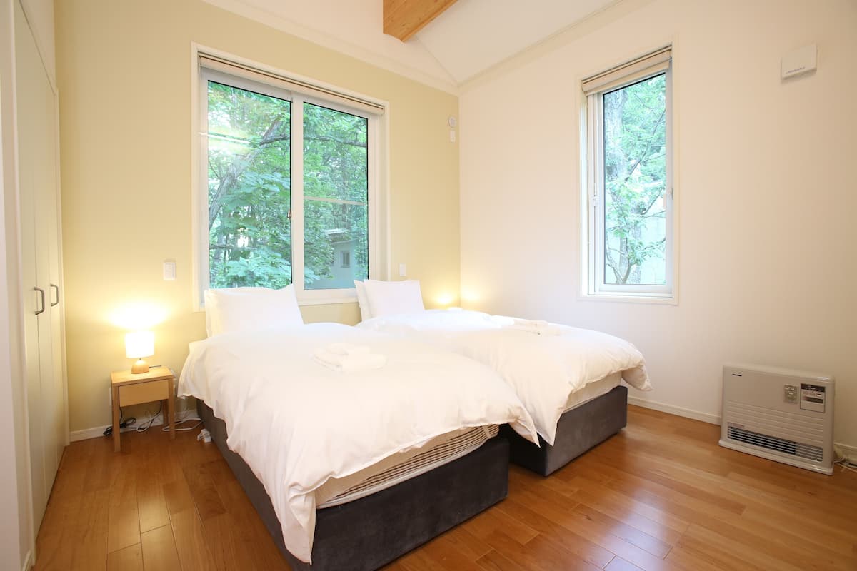 Een slaapkamer met twee bedden, een nachtkastje met lamp, een verwarming en grote ramen met uitzicht op bomen.