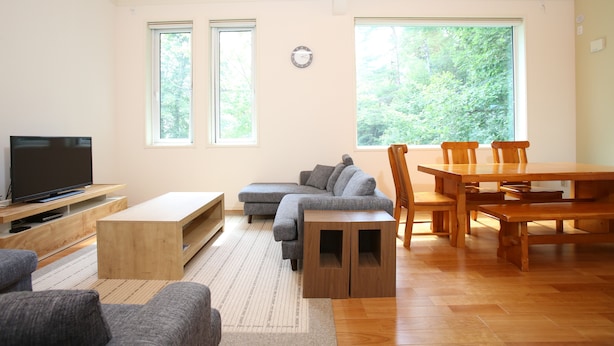 Una sala de estar con sofá, televisor, mesa de centro de madera, mesa de comedor con sillas y ventanas grandes.