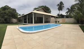 A house with a swimming pool, a covered patio, and a garden with palm trees.