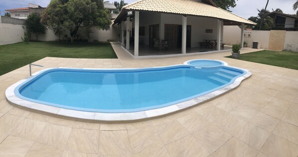 A swimming pool with a tiled deck, a covered patio area, and a grassy yard.