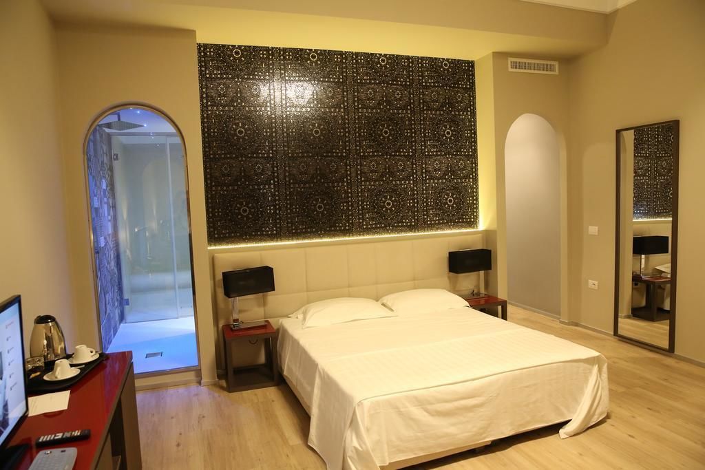 A hotel room with a large bed, two bedside tables, a desk with a computer and a lamp, a large mirror, and a decorative wall panel.