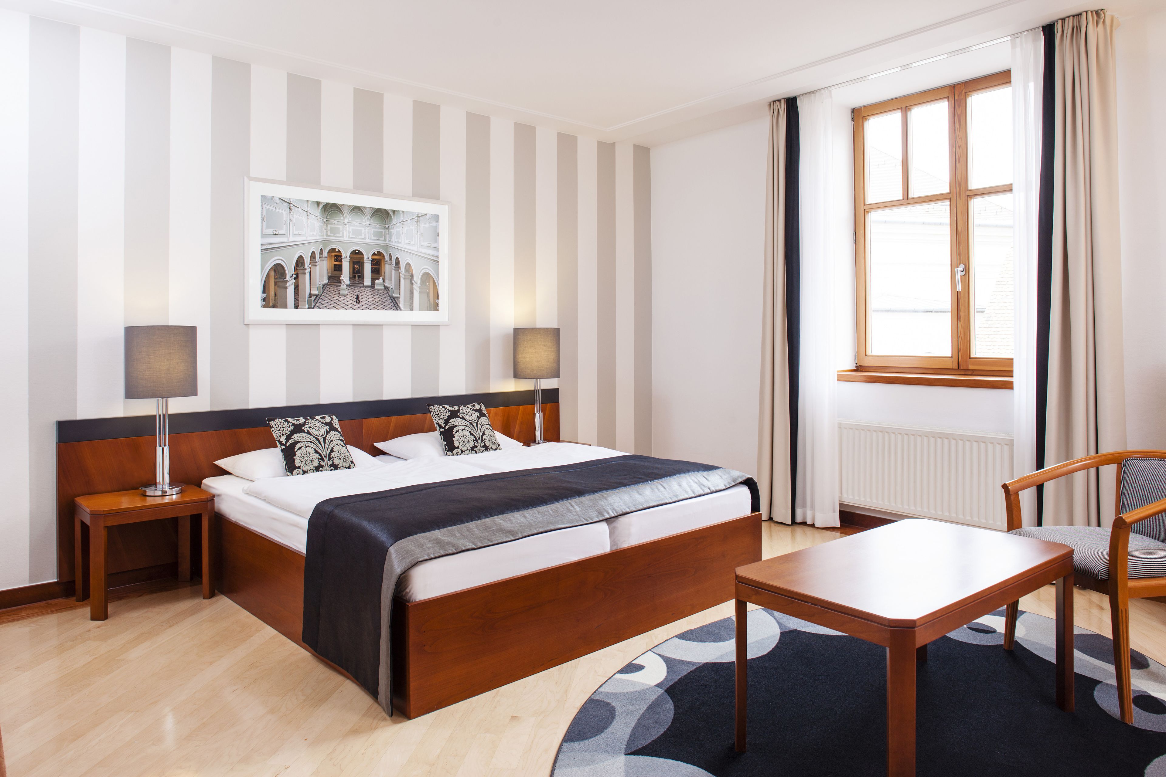 A hotel room with a large bed, two bedside tables with lamps, a wooden chair, a window with curtains, and a framed picture on the wall.