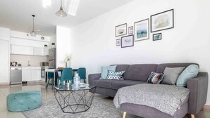 A modern living room with a grey sofa, teal ottoman, and a coffee table. There is a kitchen in the background with white cabinets and a dining area with teal chairs.