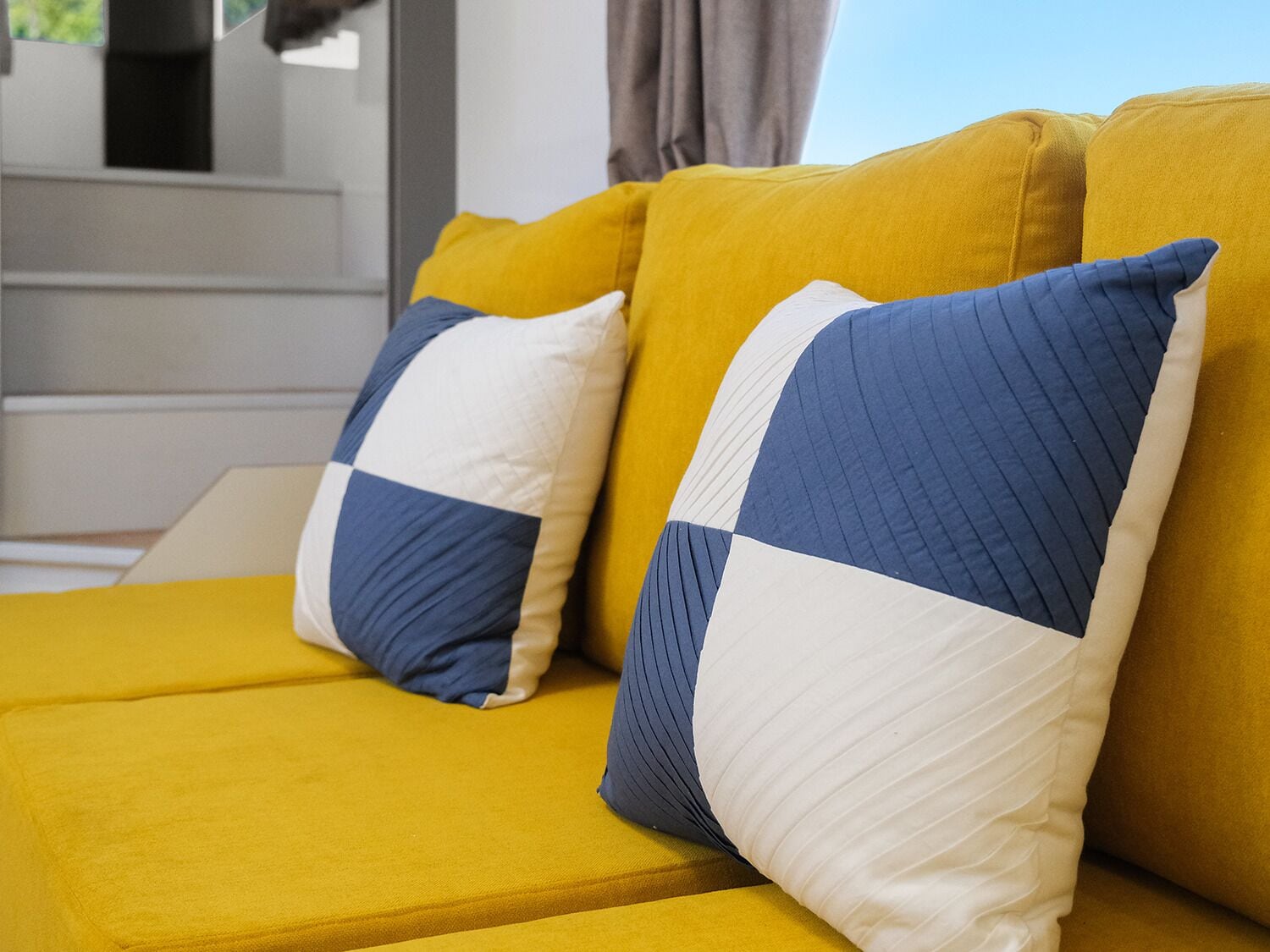 A yellow sofa with two pillows, one blue and white, and one white and blue.