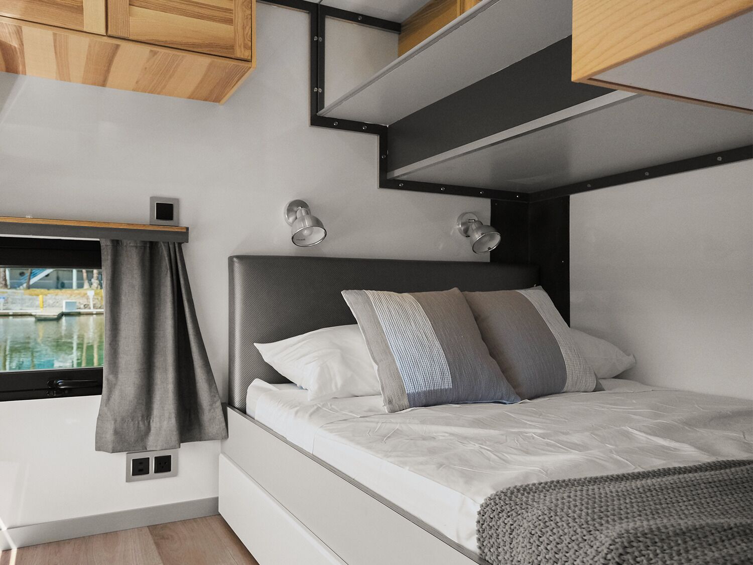 A compact bedroom with a bed, bedside lamps, a window with curtains, and a view of a body of water.