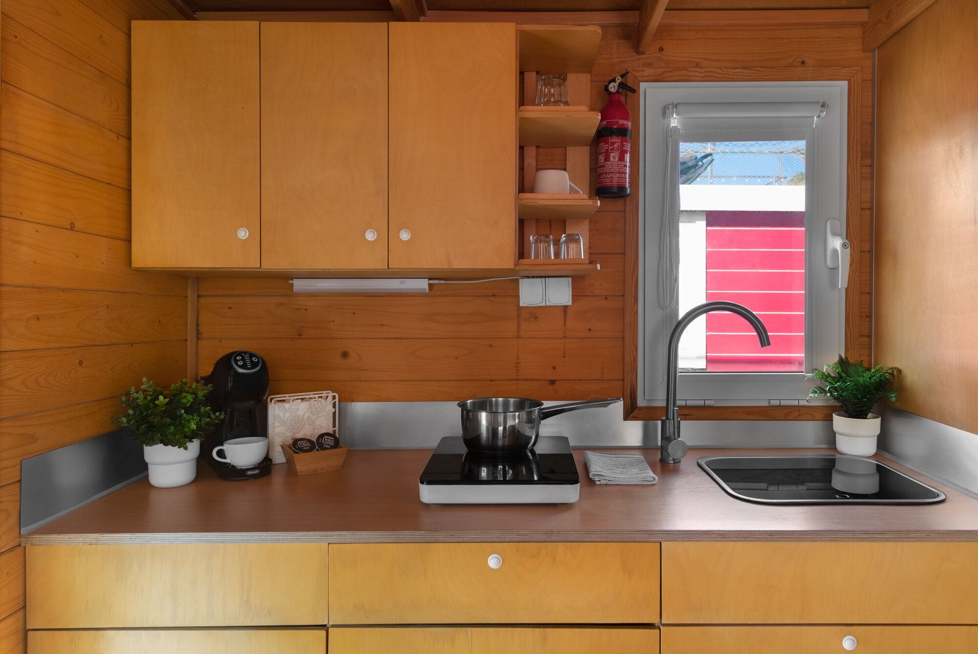 A compact kitchen with wooden cabinets, a sink, a stove, and a window with a red blind.