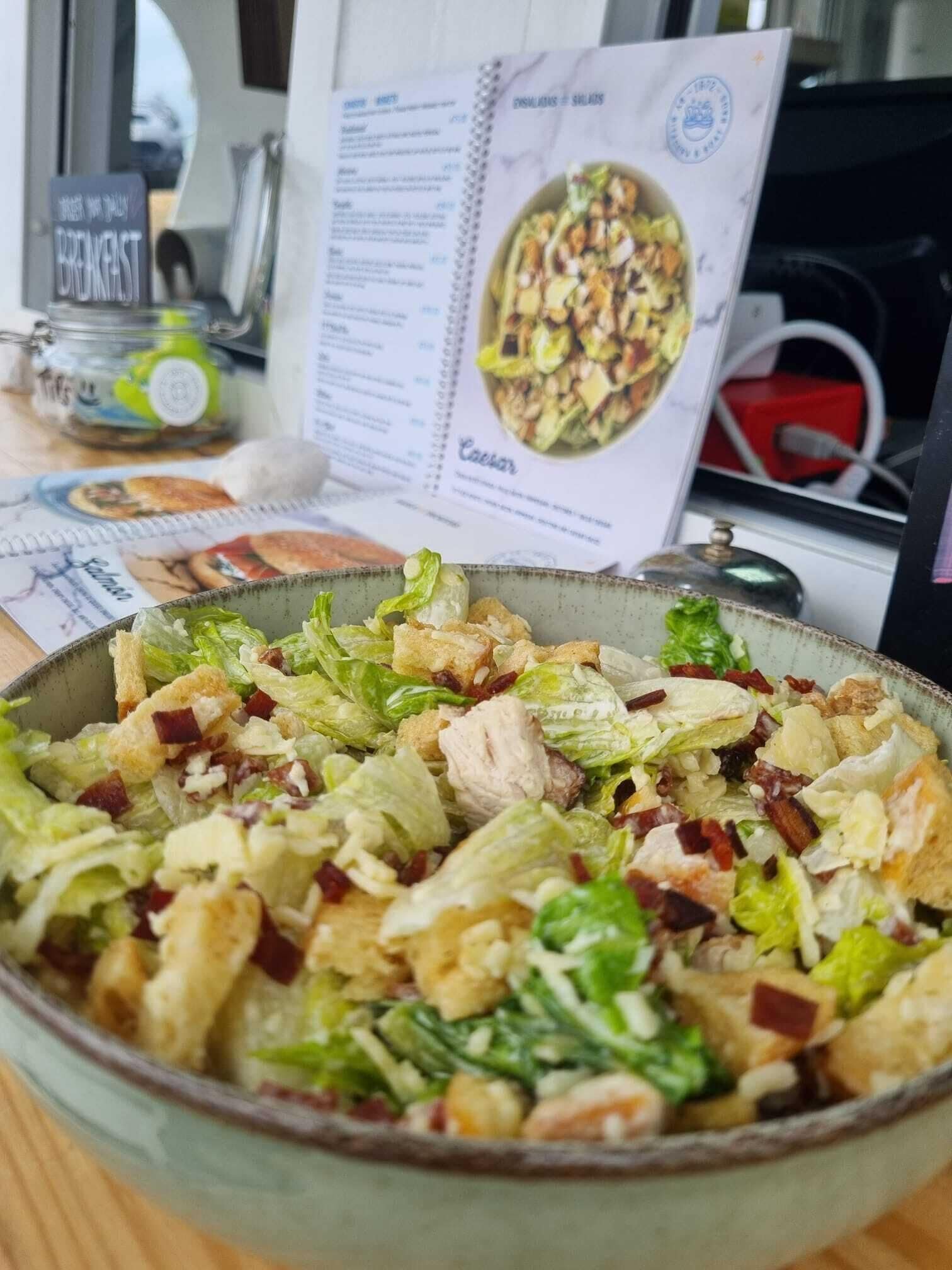 A bowl of Caesar salad with croutons and bacon, a menu with a Caesar salad picture, and a sign that says "BREAKFAST".