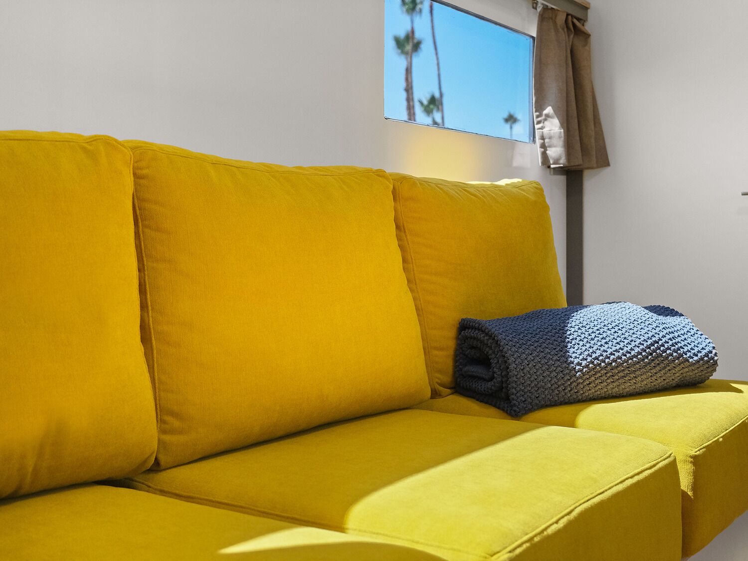 Un canapé jaune, avec une couverture pliée, et une fenêtre donnant sur des palmiers.