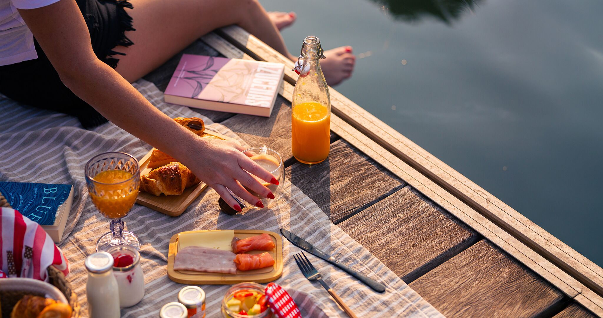 Une jetée en bois aménagée pour un pique-nique, avec une bouteille de jus d’orange, un verre de jus d’orange, un croissant, un sandwich au jambon et un livre.