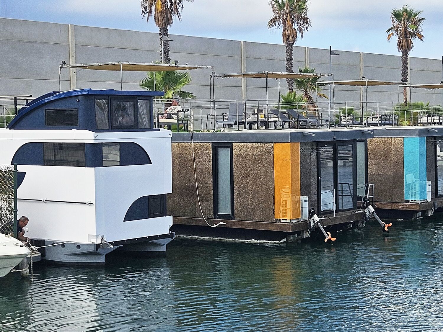 A houseboat docked at a marina with a covered deck area and a small boat nearby.