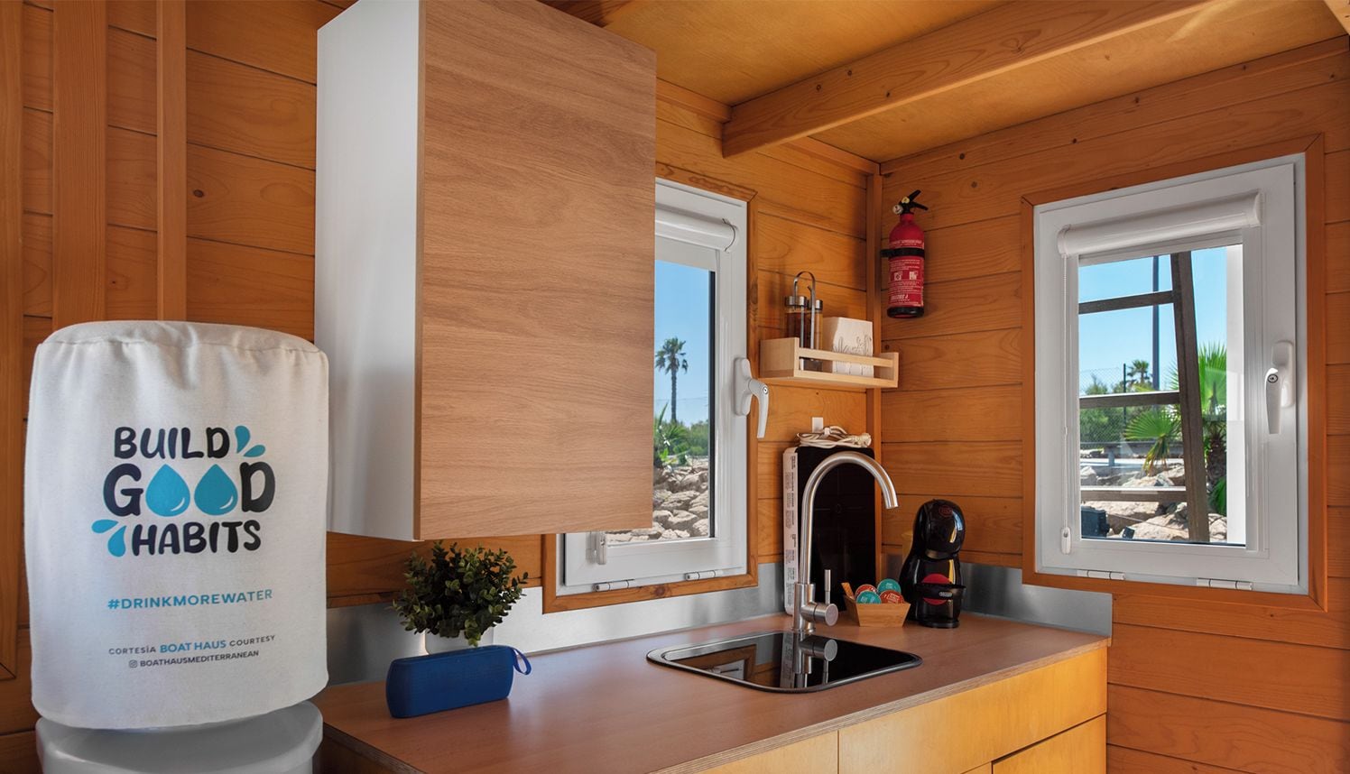 A compact kitchen with wooden walls, a sink, and two windows. A fire extinguisher is mounted on the wall.