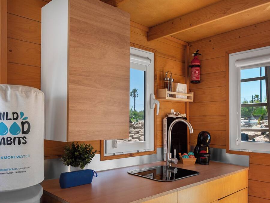 A compact kitchen with wooden walls, a sink, and two windows. A fire extinguisher is mounted on the wall.
